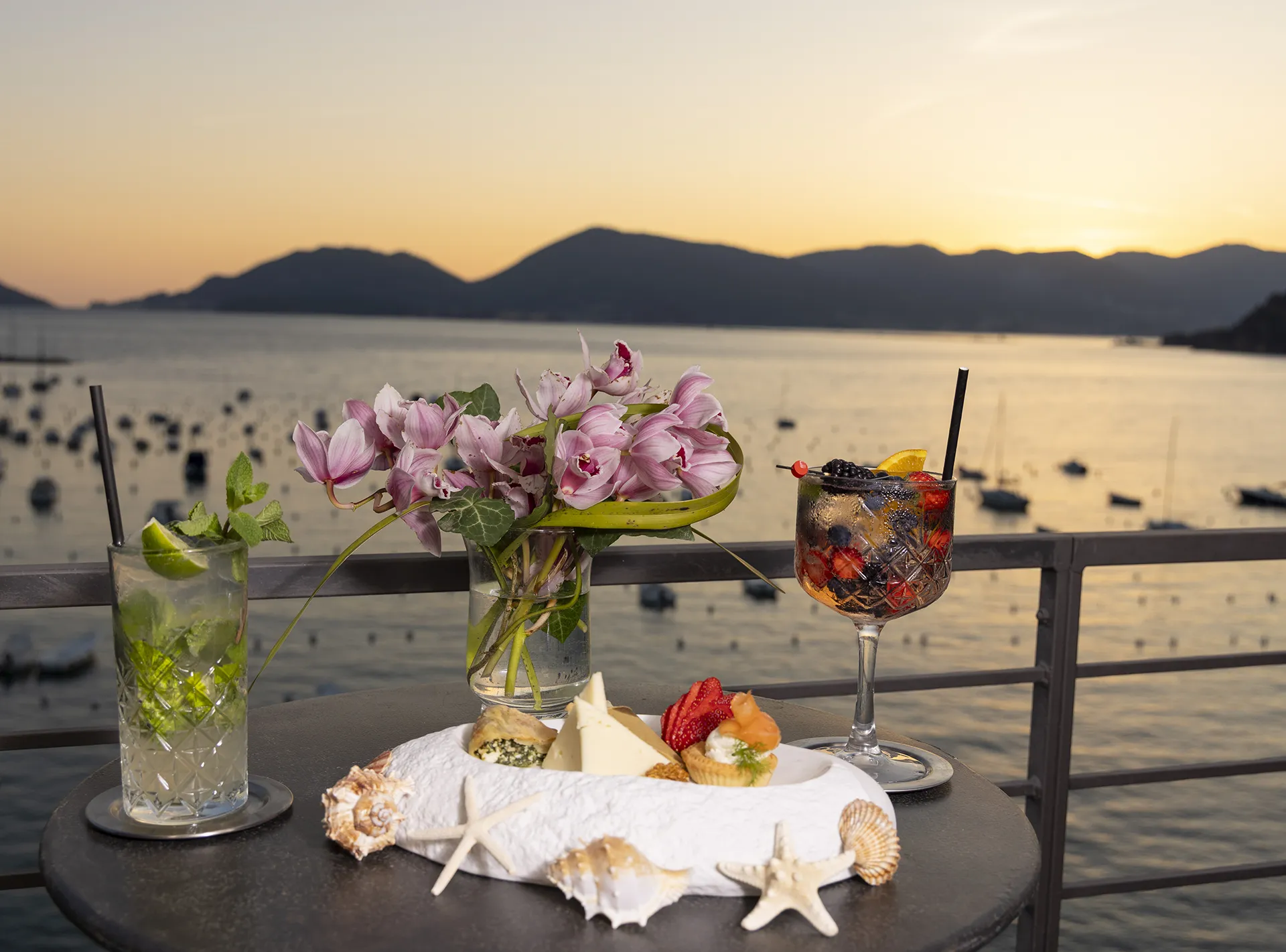 Nell'immagine, il primo piano su un tavolino su cui sono poggiati due cocktail e alcune tartine per un aperitivo presso l'Hotel Shelley e Delle Palme. Di fronte, la vista mare al tramonto.