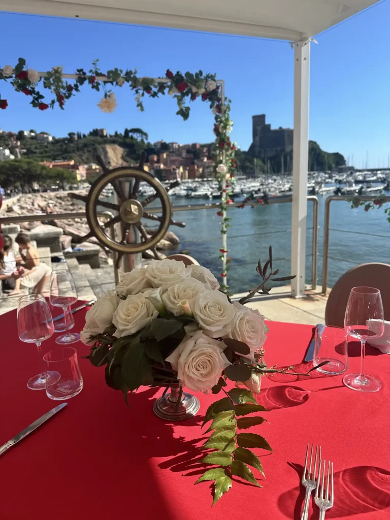 Nell'immagine, un primo piano su un tavolo con dei fiori bianchi e la tovaglia rossa, ad un matrimonio organizzato presso l'Hotel Shelley e Delle Palme. Sullo sfondo, si vede il mare e il castello di Lerici.